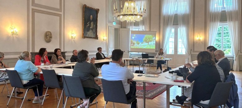 Anfang Juni war das Tourismuscluster Netzwerk zu Gast im Kurfürstlichen Schlösschen in Bad Bertrich. Dabei führte Vera Esch, Geschäftsfüh-rerin der GesundLand Vulkaneifel GmbH, durch die verschiedenen Räumlichkeiten wie den Kur-saal, den Konzertsaal und den Karlsbadsaal.In Kreisaufgestellten Tischen sitzen die Teilnehmenden und lauschen Sandra Schneemann (Tourismuscluster EMH).