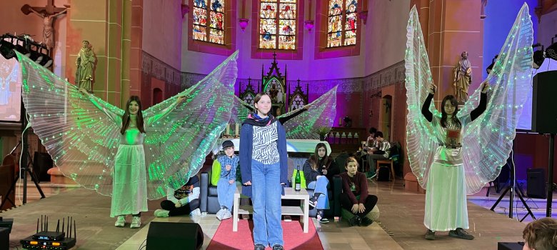 Musical-AG aus Kaisersesch gibt außergewöhnliches Gastspiel in der Nachbarschaft Musical-AG aus Kaisersesch gibt außergewöhnliches Gastspiel in der Nachbarschaft: Das Bild zeigt eine Szene des Theaterstücks, das in der Kirche in Masburg aufgeführt wird. Schülerinnen und Schüler befinden sich auf der Altarinsel, die als Bühne dient. Im Vordergrund sind drei Schülerinnen zu sehen, die große, weiße Flügel tragen. Die mittlere Schülerin wird von einem Spotlight angeleuchtet. Im Hintergrund sitzen weitere Schülerinnen und Schüler.