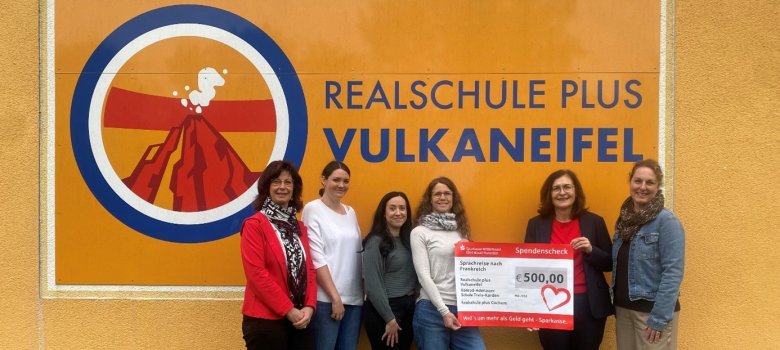 Zu sehen sind Frau Bettina Pellio und Frau Bettina Keßeler, welche die Sparkassenstiftung vertreten, sowie die stellvertretende Schulleiterin Frau Nadine Fresia und Französischlehrerin Frau Erika Heinrich der Realschule plus Vulkaneifel. Ebenfalls waren die Vertreterinnen des Födervereins der Realschule plus Vulkaneifel, Frau Katharina Theobald und Frau Eileen Enkel zu sehen. Alle zusammen stehen vor dem Logo der Realschule plus Vulkaneifel, wo der Scheck von 500 Euro übergeben wird.