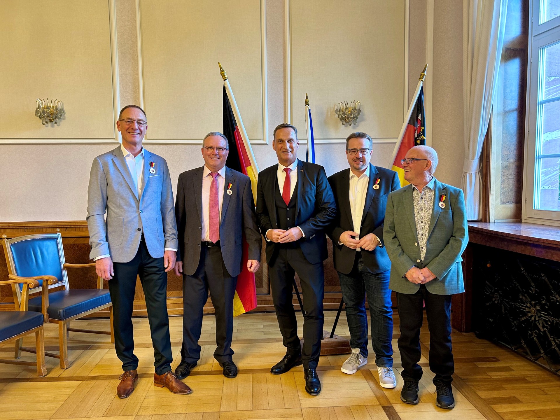 Auf dem Foto sind von links folgende Personen zu sehen: Günter Groß, Berthold Berenz, Joachim Rodenkirch, Sebastian Seifert und Karl-Heinz Hilgenberg. Sie stehen in einem Saal mit Holzboden vor einem Aufsteller mit drei Flaggen (Deutschland, unbekannt, Rheinland-Pfalz) in einer Reihe.