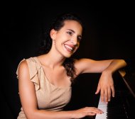 Melisa Cingolani Das Bild zeigt die Pianistin Melisa Cingolani. Sie sitzt an einem Flügel, ein arm liegt lässig auf dem Instrument, die Hand des anderen Arms liegt auf den Tasten. Der Hintergrund ist schwarz. Melisa Cingolani trägt ein beiges Kleid und lächelt.