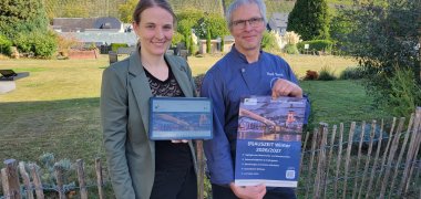 (R)AUSZEIT 2026/27 – Zahlreiche Winter-Angebote für Gäste und Einheimische im Land-kreis Rudi Burch und Steffi Schmitz vom Hotel „Zur Marienburg“ stehen nebeneinander und halten ein Plakat bzw ein Tablet in den Händen, auf dem Werbung für "(R)AUSZEIT" zu sehen ist. Hinter ihnen sieht man eine Wiese sowie ganz in der Ferne den Berg mit der Marienburg.