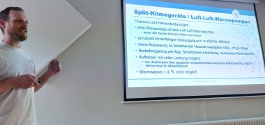 Sonne nutzen und Zuhause kühlen: Große Re-sonanz bei Info-Veranstaltung in Treis-Karden Das Bild zeigt einen Mann bei der Präsentation seines Vortrags. Er steht links im Bild vor einer Wand und zeigt zu einer Leinwand, auf der Text zu sehen ist.
