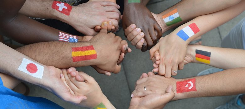 Menschen mit verschiedenen Hautfarben haben unterschiedlichen Flaggen auf ihren Armen gemalt und stehen miteinander im Kreis und halten sich die Hände.