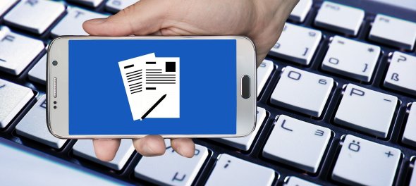 Eine Hand umfasst ein Smartphone vor einer Tastatur. Auf dem Bildschirm des Smartphones ist ein Formular zu erahnen. Eine Hand umfasst ein Smartphone vor einer Tastatur. Auf dem Bildschirm des Smartphones ist ein Formular zu erahnen.
