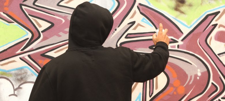 Dargestellt ist eine Person im schwarzen Kapuzenshirt von hinten der ein Graffiti sprüht.