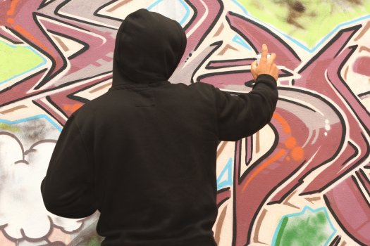 Dargestellt ist eine Person im schwarzen Kapuzenshirt von hinten der ein Graffiti sprüht.