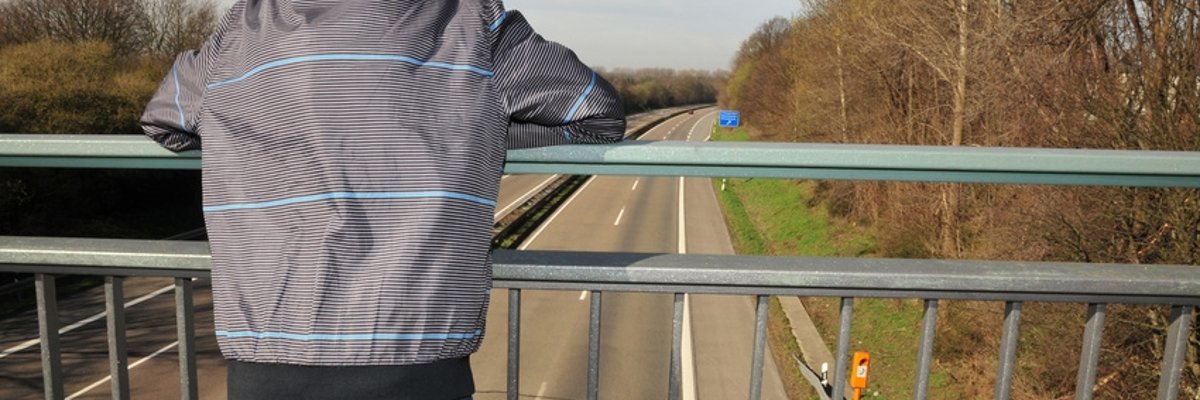 Dargestellt ist ein junger Mensch in einer karierten Jacke von hinter, der auf einer Autobahnbrücke steht. Dargestellt ist ein junger Mensch in einer karierten Jacke von hinter, der auf einer Autobahnbrücke steht.