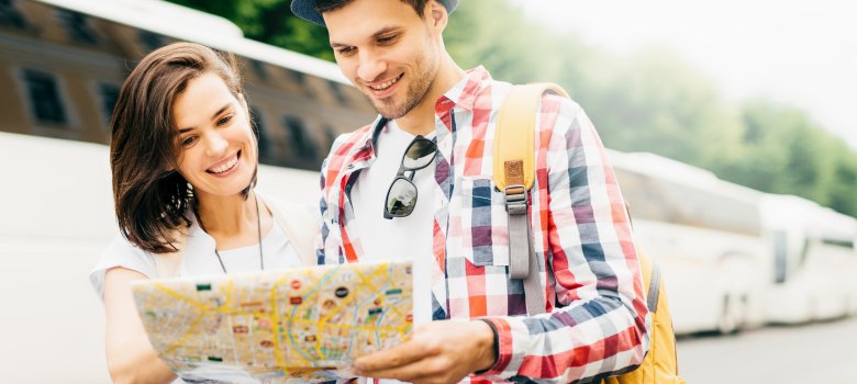 Junger Mann und junge Frau stehen zusammen und schauen auf einen Plan. Im Hintergrund stehen Busse.