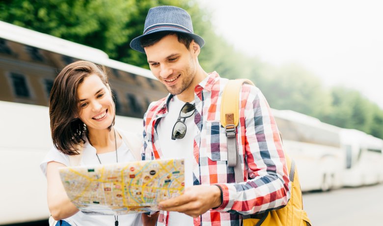 Junger Mann und junge Frau stehen zusammen und schauen auf einen Plan. Im Hintergrund stehen Busse.