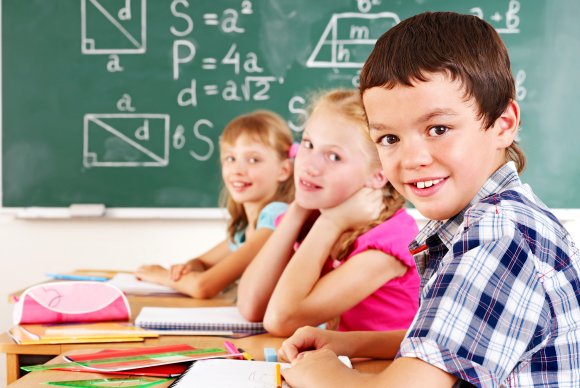 Drei Kinder, ein Junge und zwei Mädchen, blicken über Ihren Schreibtisch in der Klasse hinweg in die Kamera. Im Hintergrund ist eine Tafel mit mathematischen Formeln zu sehen.