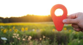 A hand is holding a red location marker in the sunset background. The concept of tourism and travel. Navigation and exploration. Destination. Holiday, vacation. Buying building land. Selective focus